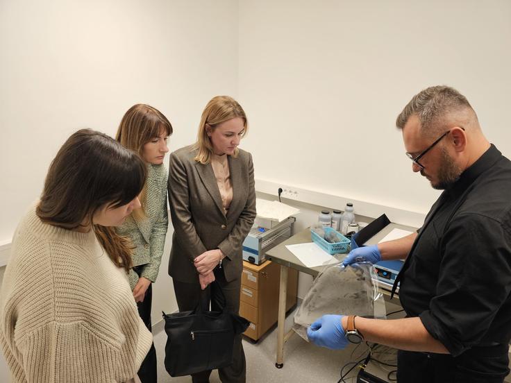 A man in gloves demonstrates an item to three women in a lab.