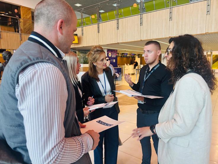 A small group of aviation security professionals stand in discussion inside Chișinău International Airport during an OSCE–ICAO training activity. Participants wearing accreditation badges review documents and exchange ideas in a bright airport terminal, reflecting the practical and collaborative nature of the Aviation Security (AVSEC) National Inspectors Course in Moldova.