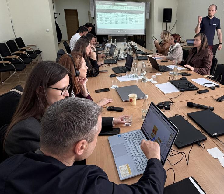 People seated at a conference table with laptops, engaging in discussion. Presentation on a screen in the background.