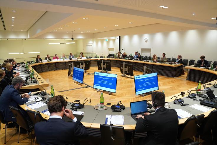 Conference room with people seated around a large, U-shaped table, screens displaying information at the center.
