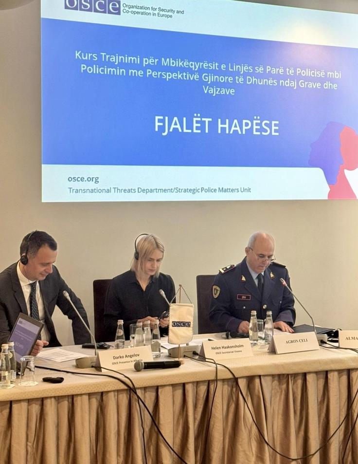 Three people sit at a conference table with microphones and documents, with a presentation screen behind them.