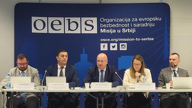 Five people in suits sit at a conference table against a blue backdrop with logos and text in Serbian.