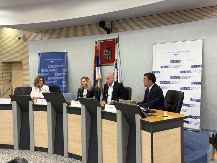 Four people sit at a conference panel table with microphones, flags, and banners in the background.