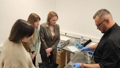 A man in gloves demonstrates an item to three women in a lab.