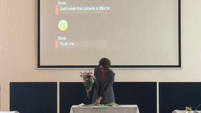 A person standing at a table with a bouquet of flowers, under a projection screen displaying text.