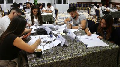 Students sitting at tables in a classroom, concentrating on writing tasks with papers and pens.