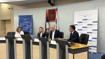 Four people sit at a conference panel table with microphones, flags, and banners in the background.