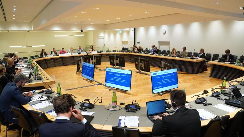 Conference room with people seated around a large, U-shaped table, screens displaying information at the center.